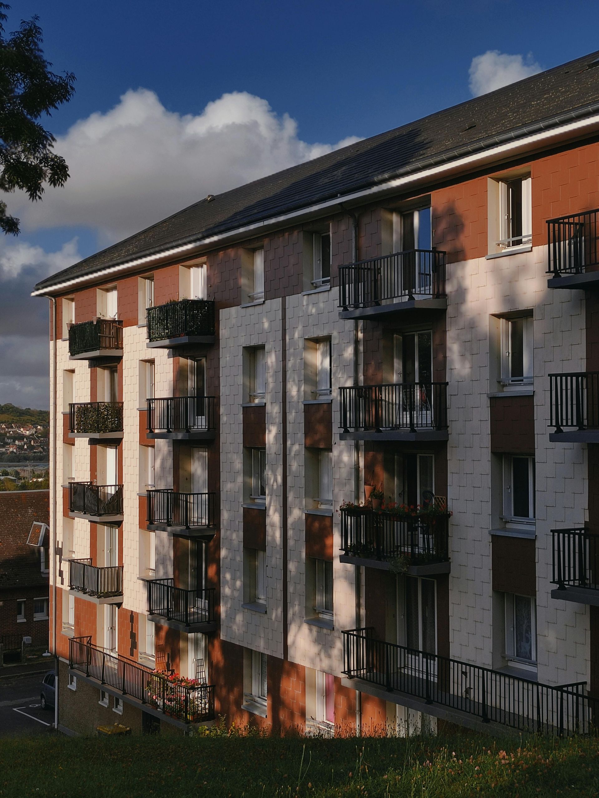 Apartment building with balconies, brown and beige facade, dark roof, against a blue sky with clouds.