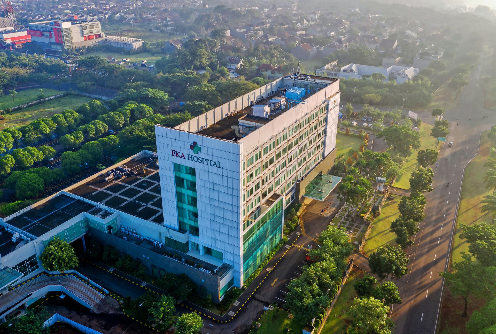 Aerial view of a modern hospital building with green accents, trees, and a roadway.