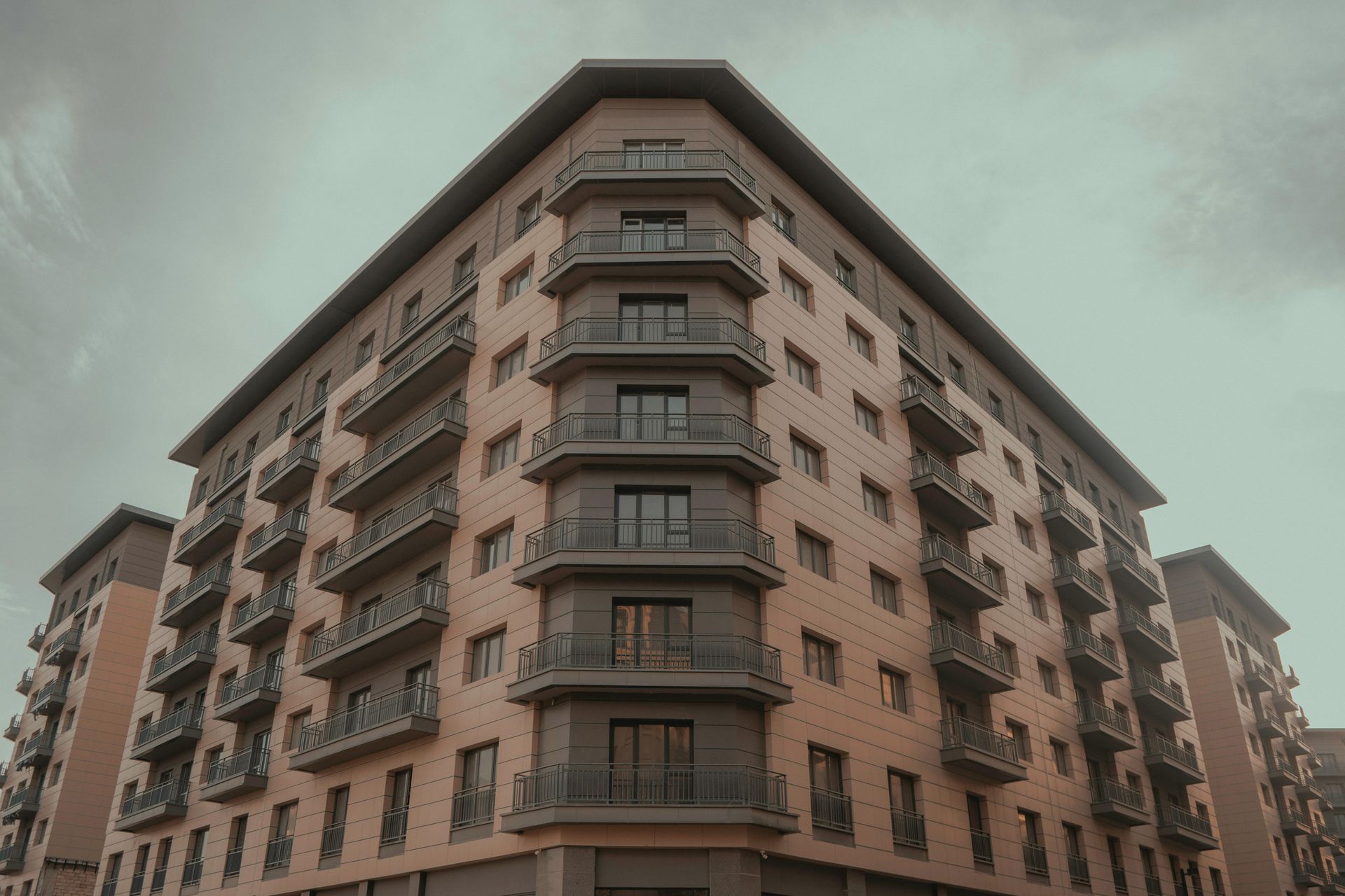 Multi-story apartment building with balconies, tan brick facade, overcast sky.