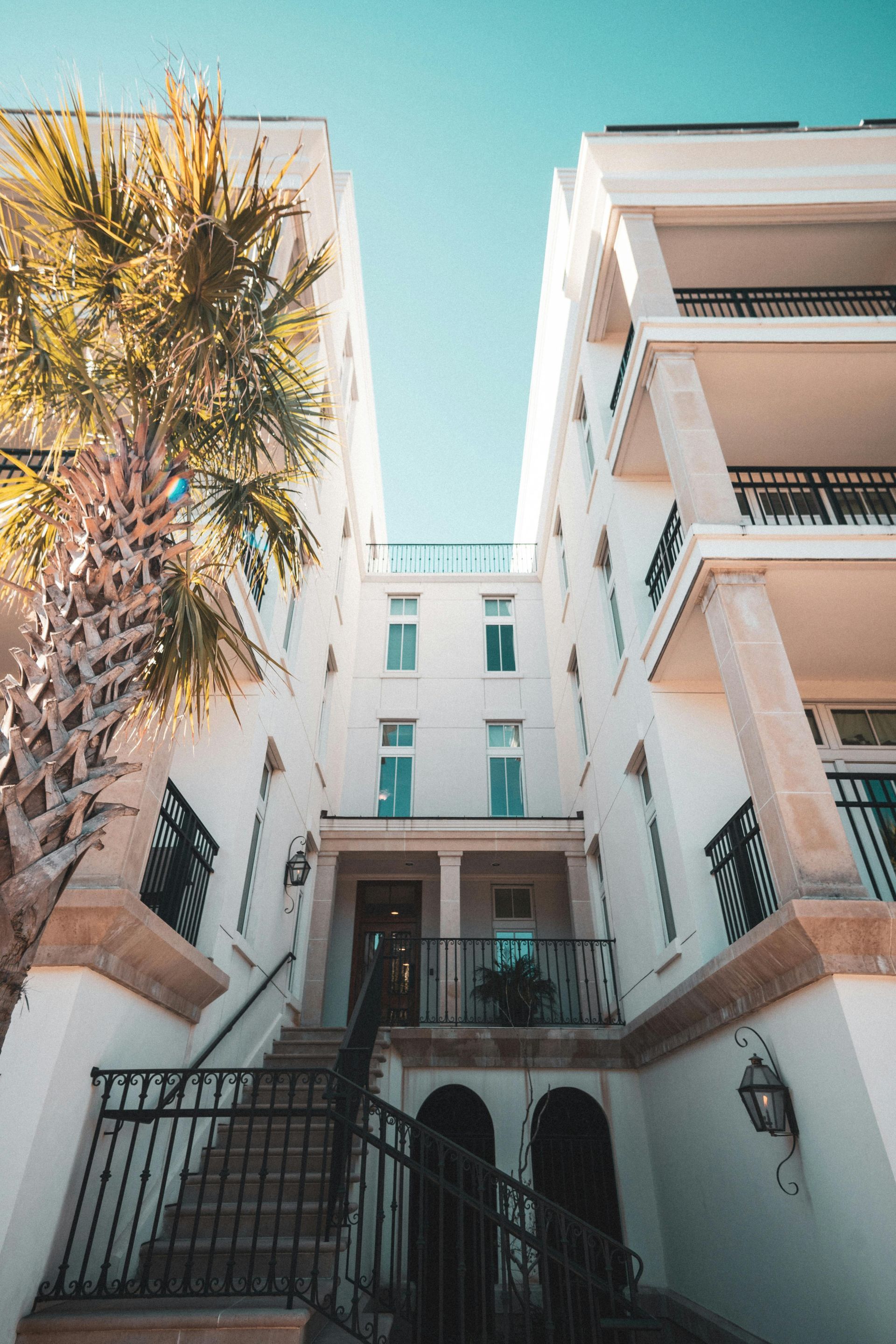 White building with balconies and stairs, palm tree on left, blue sky.