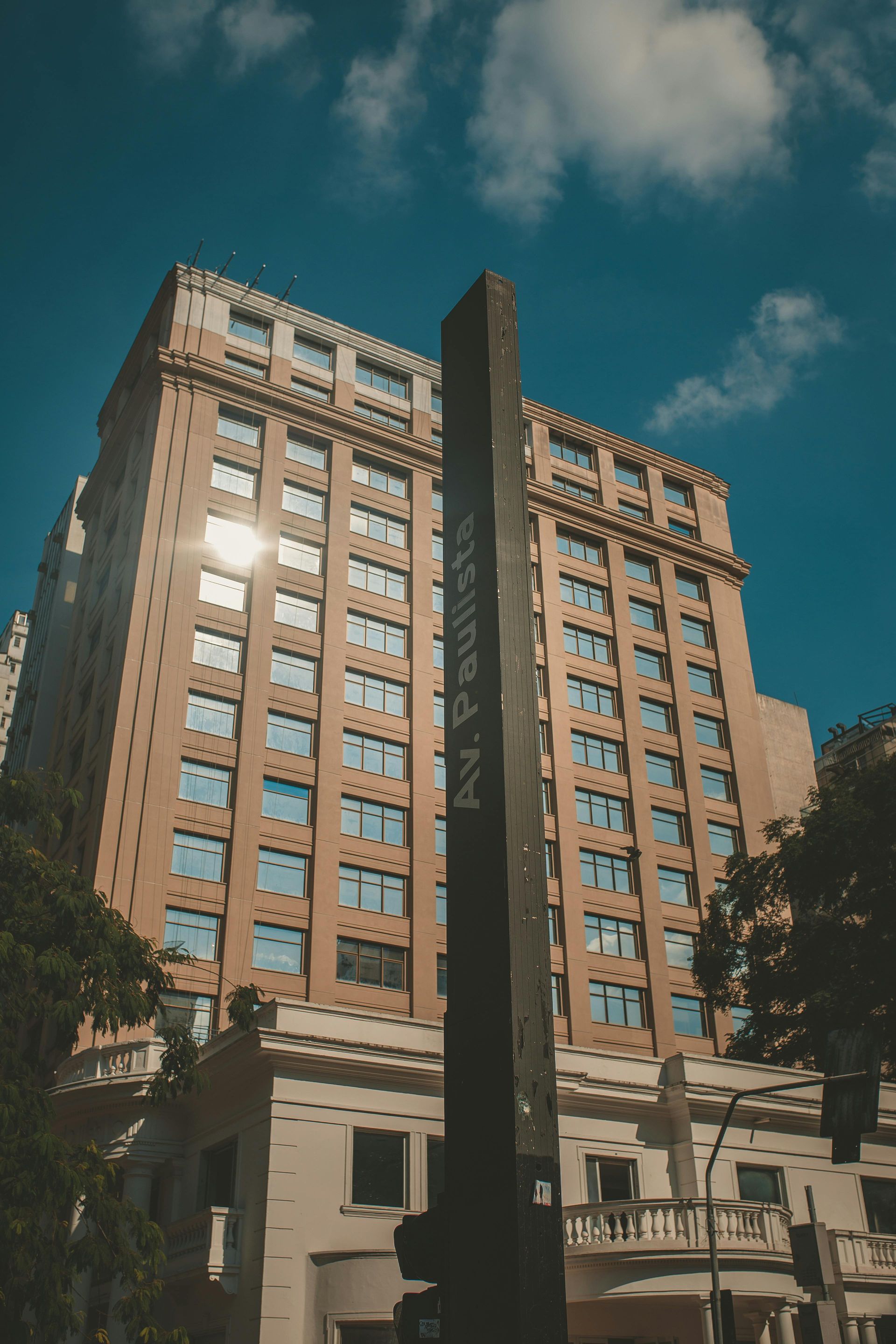 Tall building with many windows behind a dark obelisk against a bright blue sky.