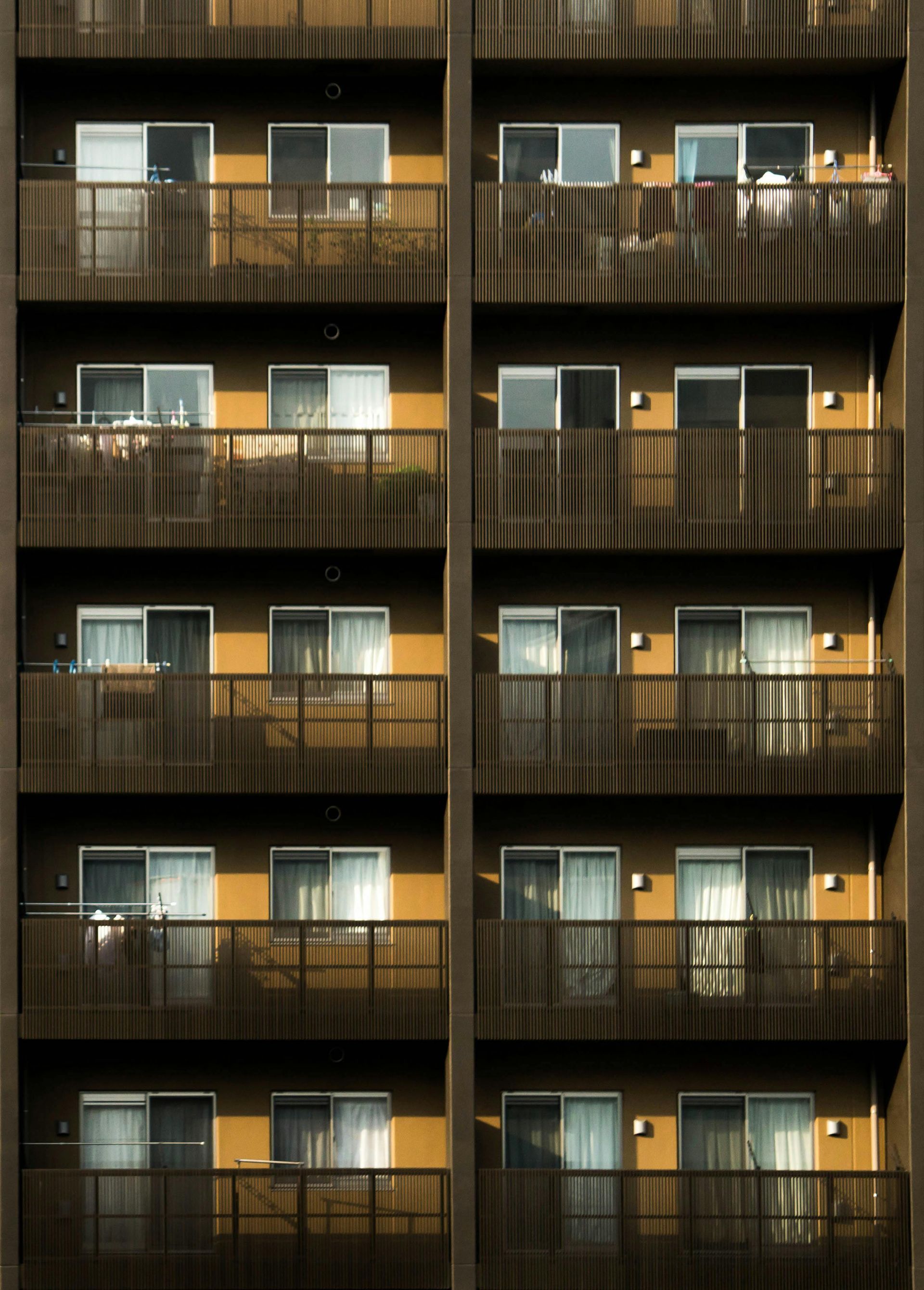 Apartment building exterior with multiple rows of balconies. Brown railing, yellow walls, and windows.