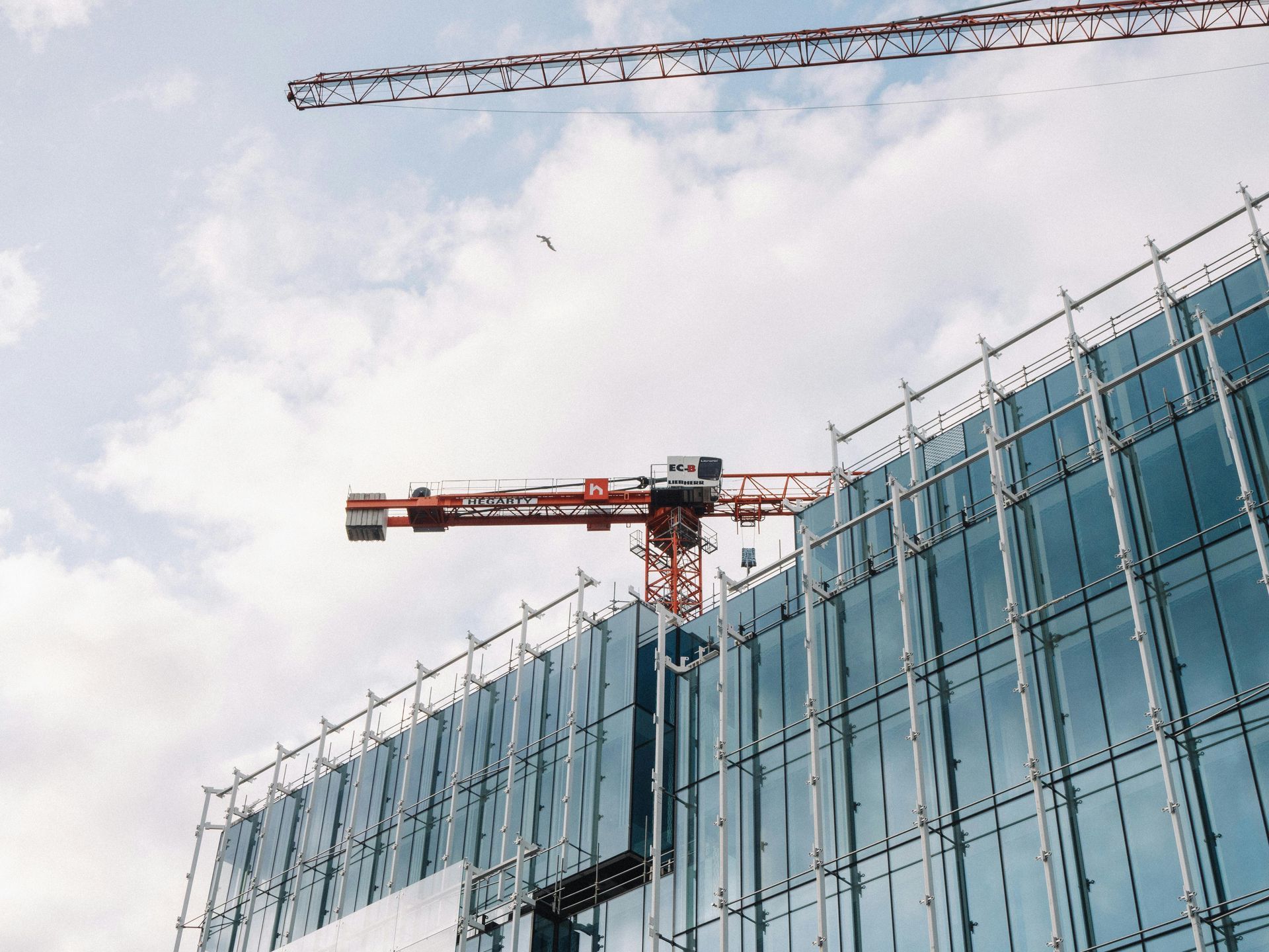 Construction site with cranes, scaffolding, and a glass-walled building under construction against a cloudy sky.