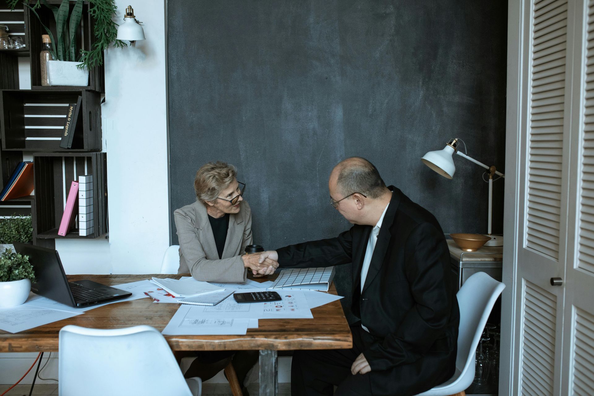 Two businesspeople shake hands at a desk, documents and laptop present, in an office setting.Check icon
