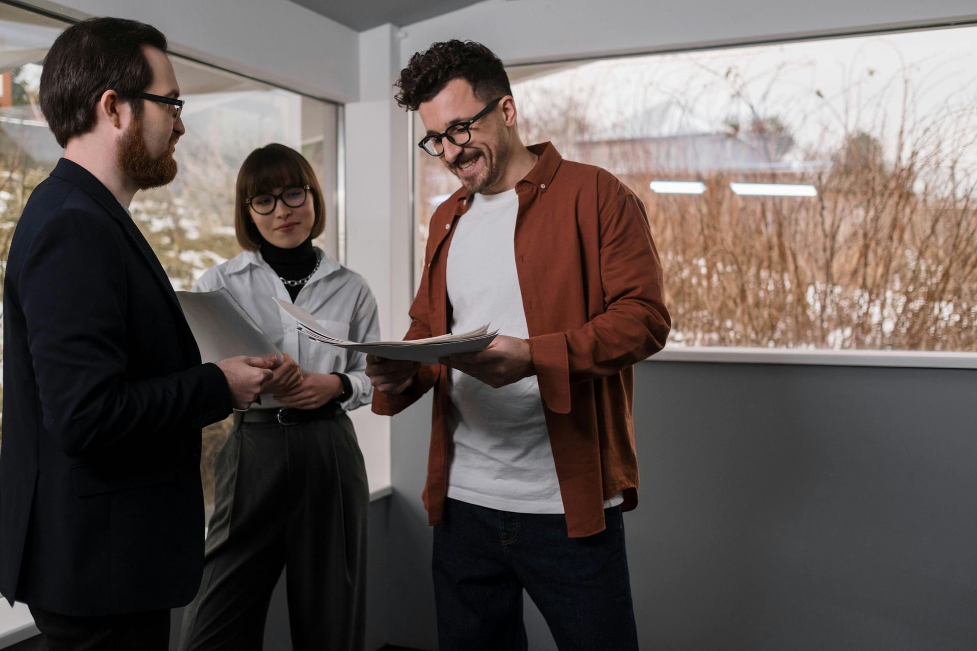 Three people looking at papers in an office near a window; one man smiles, two wear glasses.