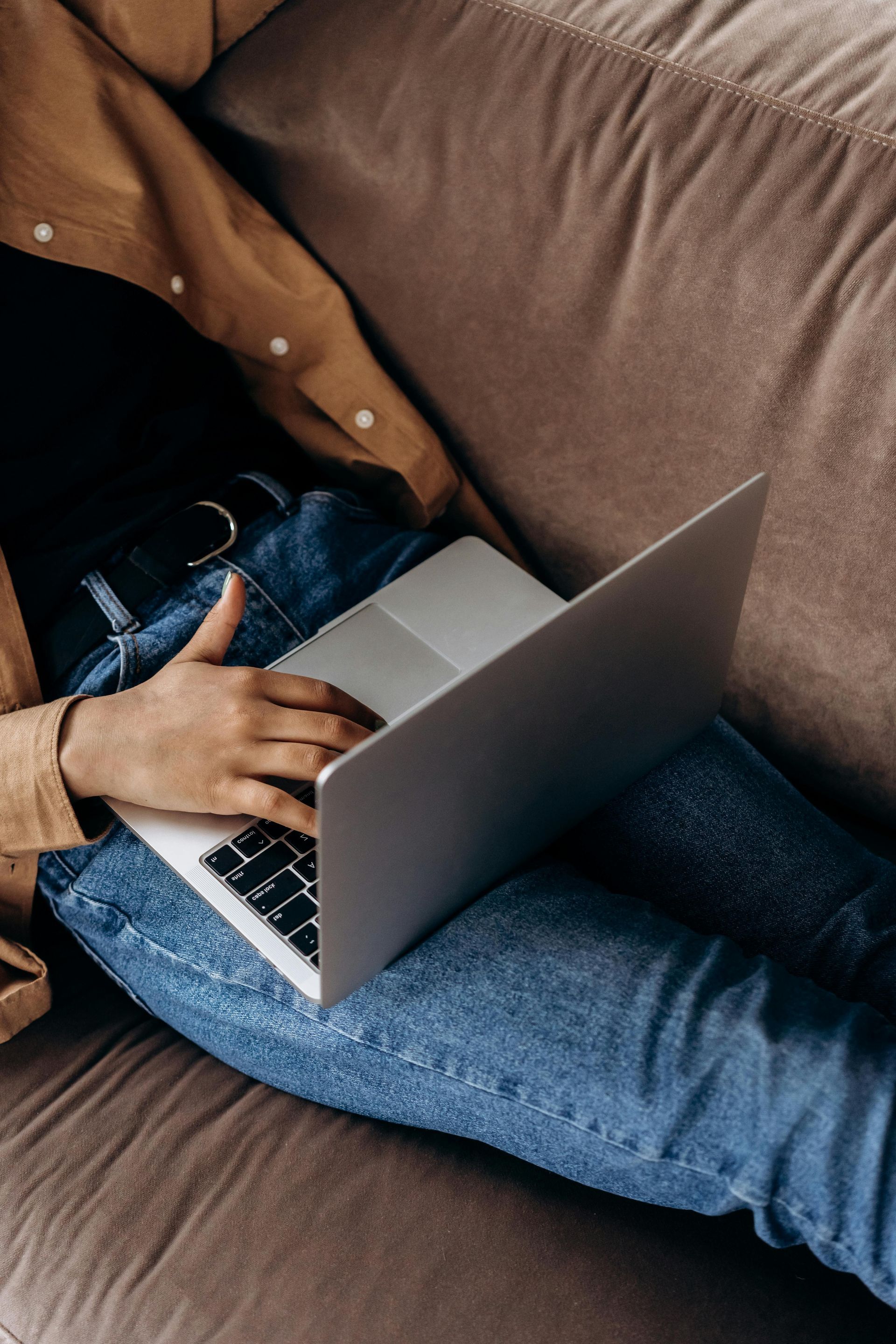 Person on a brown couch, using a laptop on their lap, wearing a brown jacket and jeans.