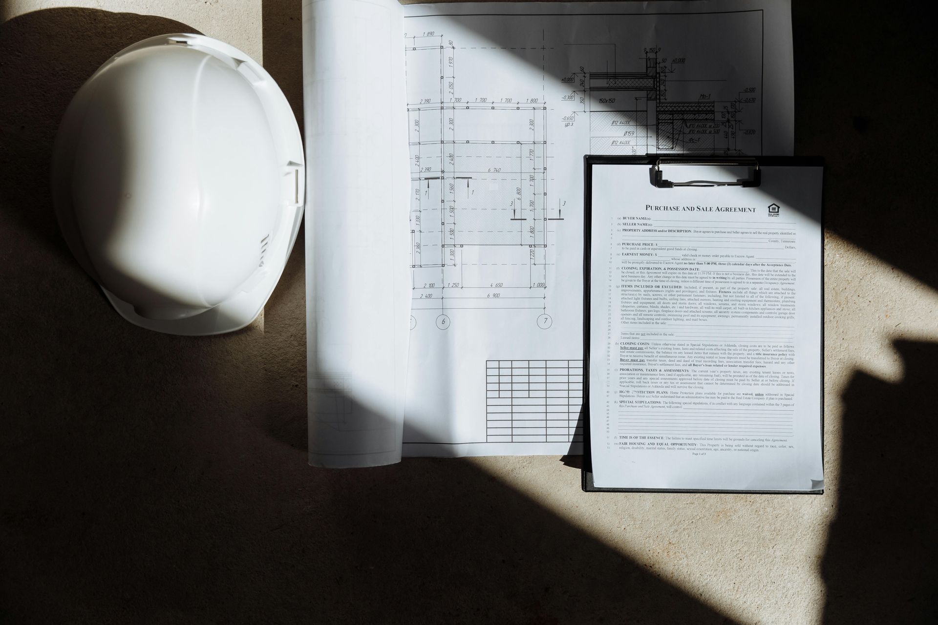 White hard hat, rolled blueprints, and clipboard with document on a concrete surface, lit by sunlight.