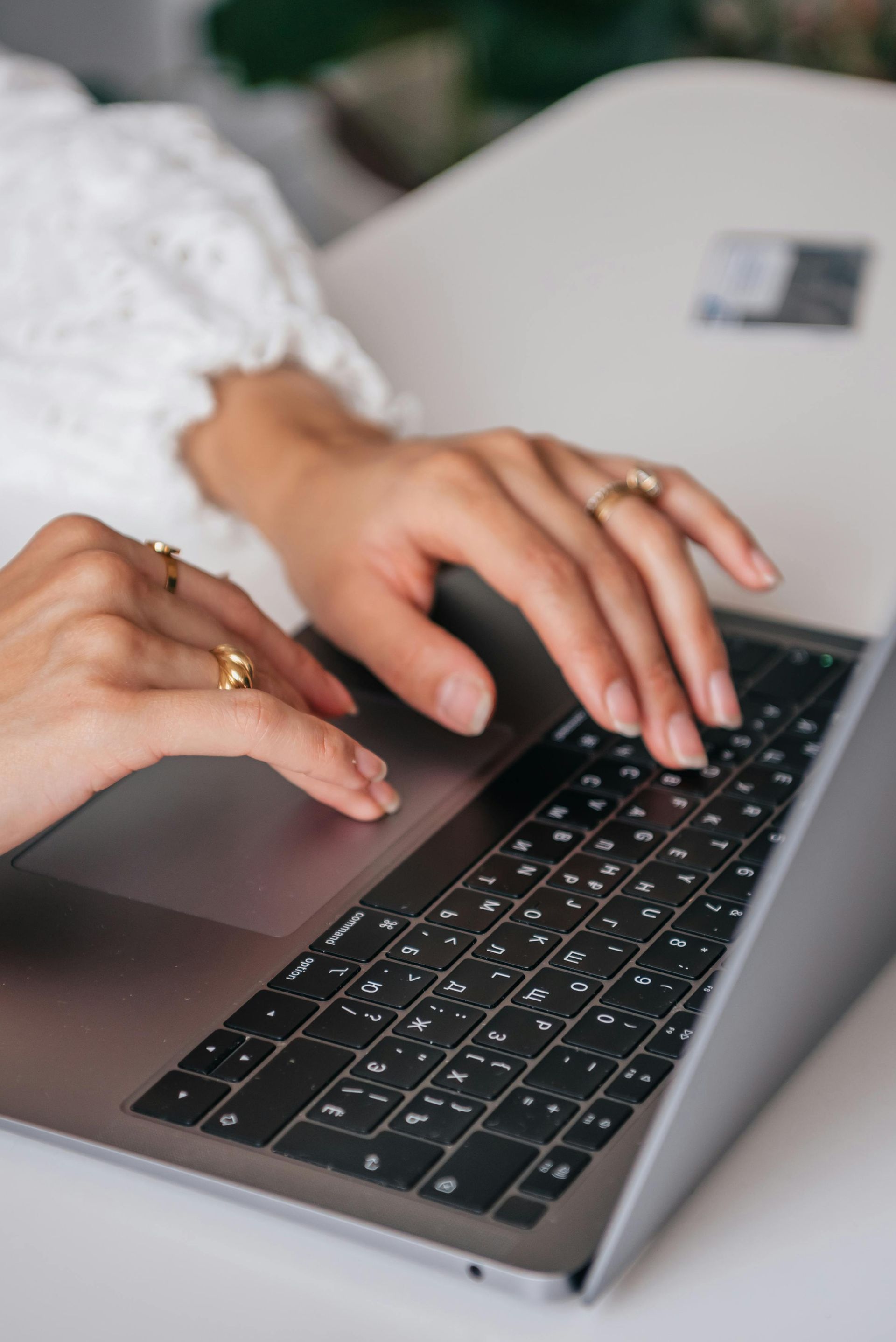 Hands typing on a laptop keyboard, wearing gold rings, with a white-sleeved top.