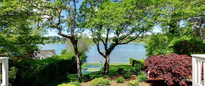 A scenic view of a lake from a balcony, framed by lush trees, a red Japanese maple, and a backyard tennis court below.