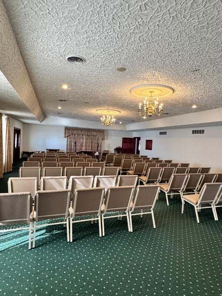 An empty auditorium with rows of beige chairs on a green carpet, under a textured ceiling and chandeliers.