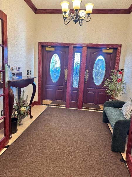 Entrance foyer with double doors, oval glass windows, dark wood trim, brown carpet, chandelier, and a small sofa.