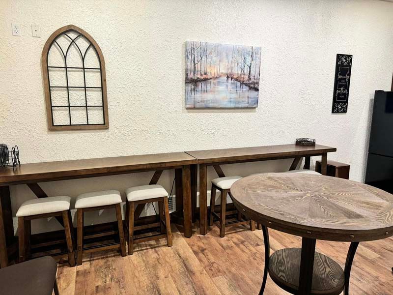 Room with wooden tables, stools, and arched window decor. Painting and decorative sign on wall.