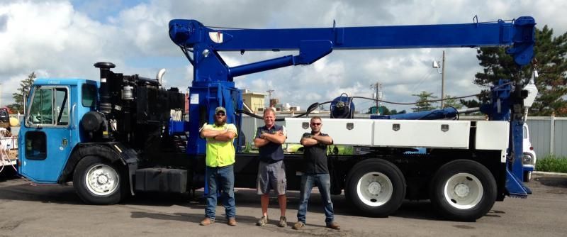 Three men stand in front of a blue crane truck with arms crossed. White cloud-filled sky.