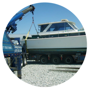 A blue tow truck lifts a white boat onto a trailer on a gravel surface. Sunny day.