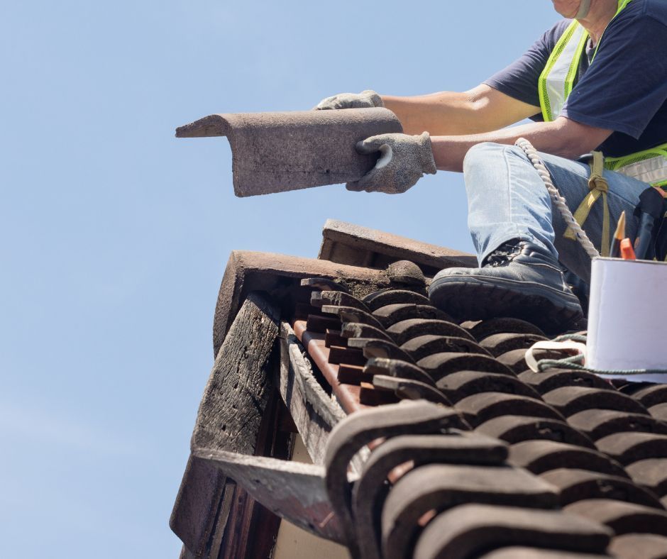 A man is sitting on top of a roof holding a tile.