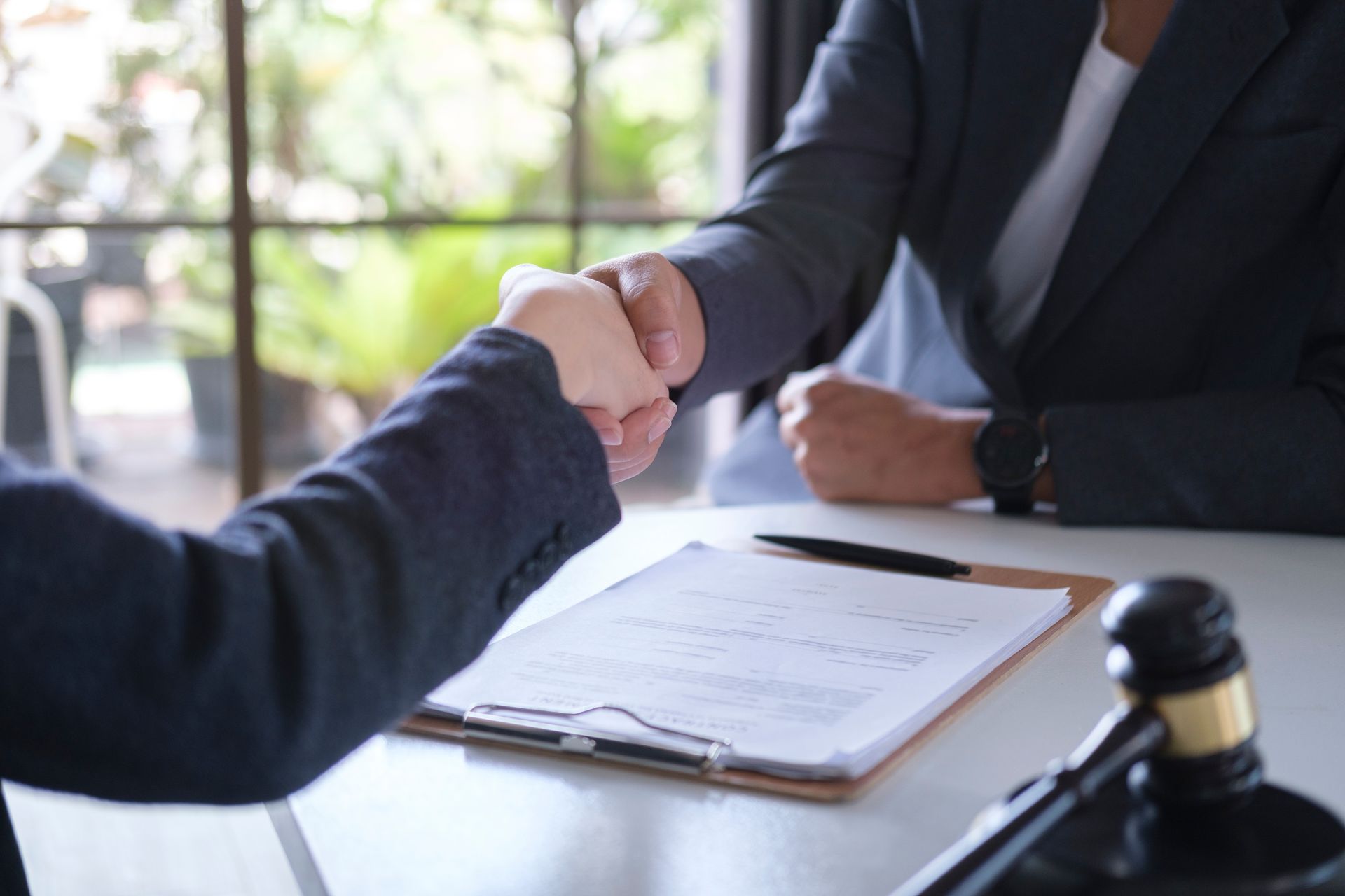 Two people in suits shaking hands over a document and gavel on a desk.