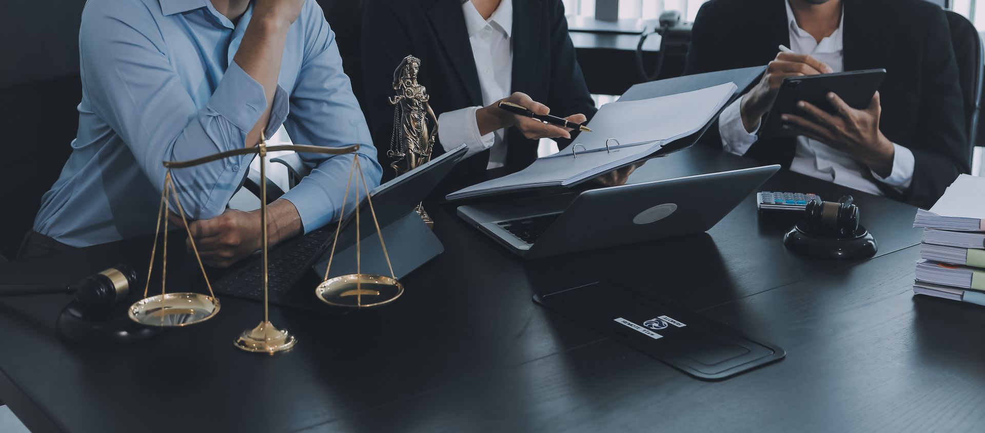 Lawyers around a table, scales of justice in the foreground. They are working on laptops and tablets, discussing documents.