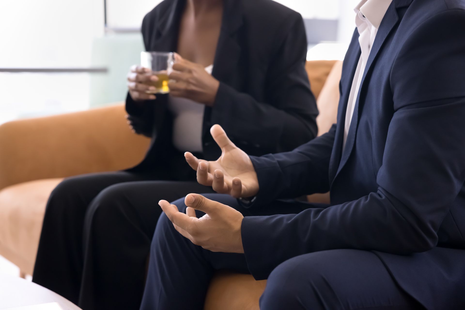 Man in suit gestures while talking; woman in background holds a drink. They are seated on a sofa.