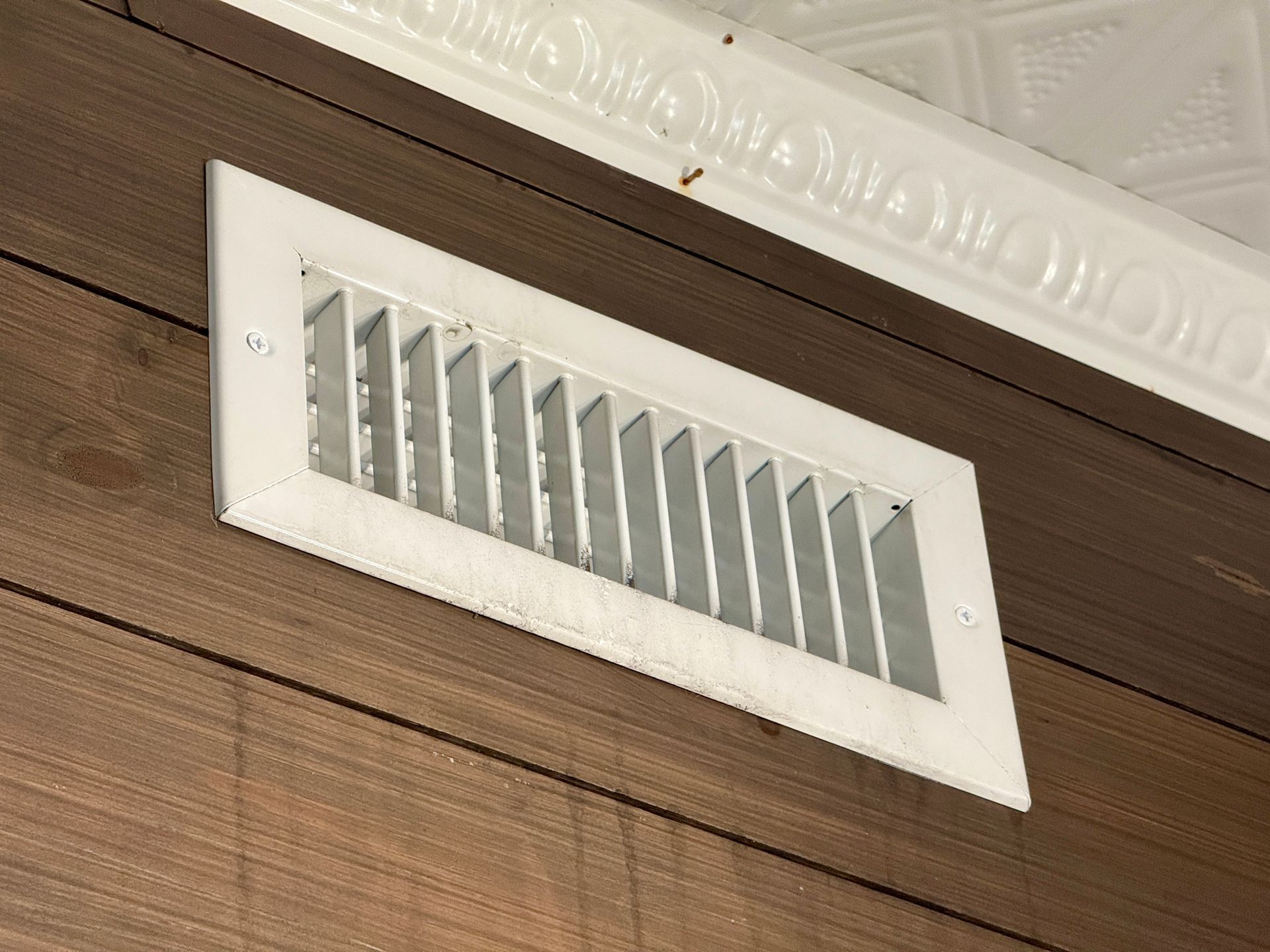 White rectangular air vent on brown paneled wall, near decorative ceiling trim.