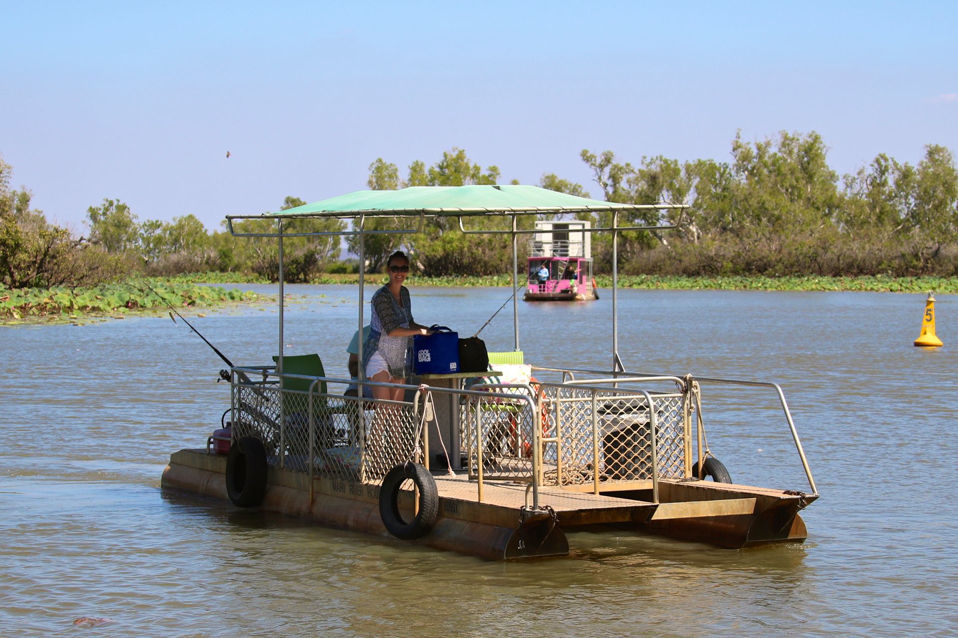 A small pontoon boat with passengers travels on a waterway. Trees and blue sky form the background.