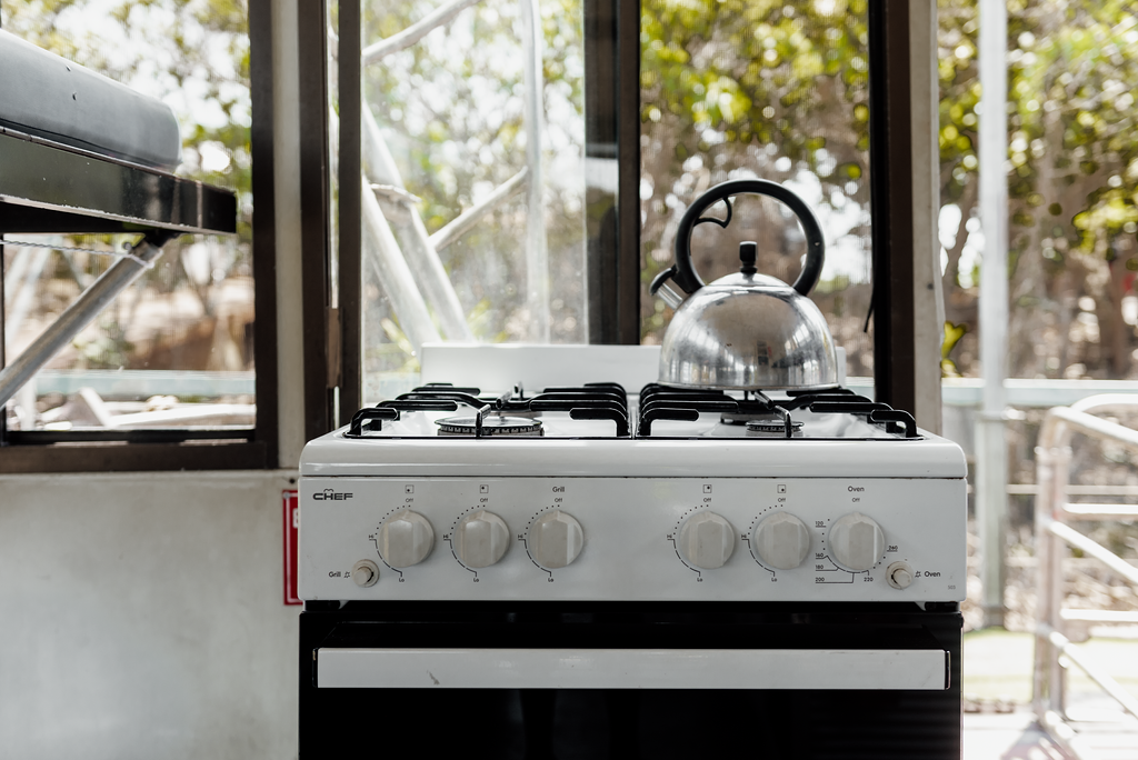 White gas stove with a kettle on top, near a window with trees visible.