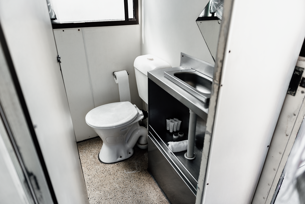 Small, basic bathroom with toilet, sink, and toilet paper. White walls, grey floor.