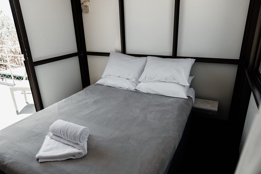 Small bedroom with bed, white pillows, gray blanket, folded towels, and dark wood trim.