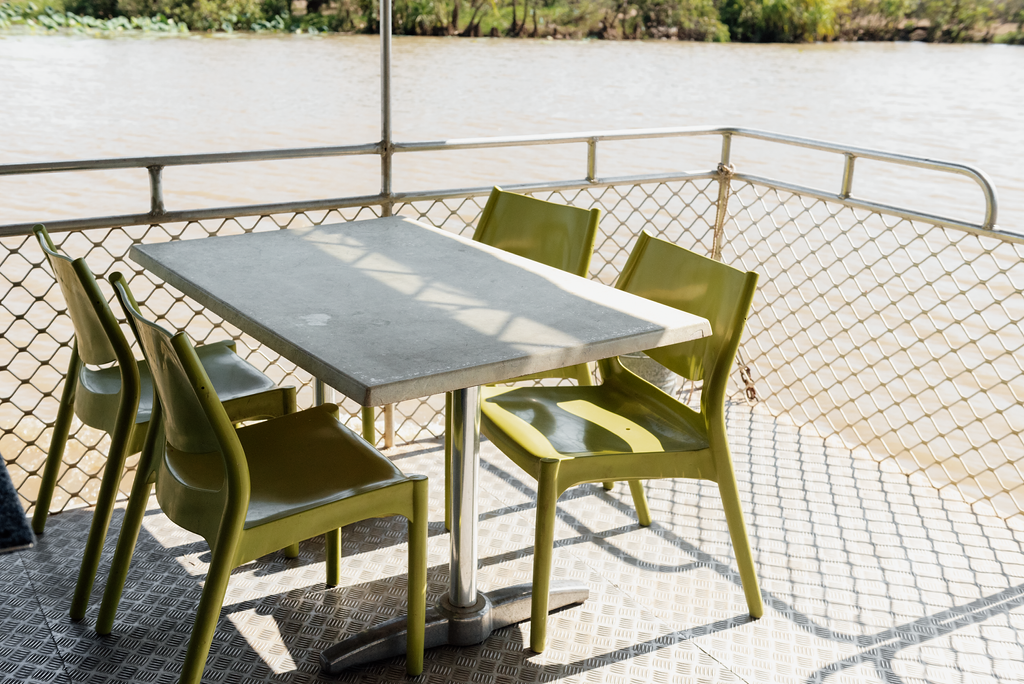 Table and green chairs on a boat deck, with a river in the background.
