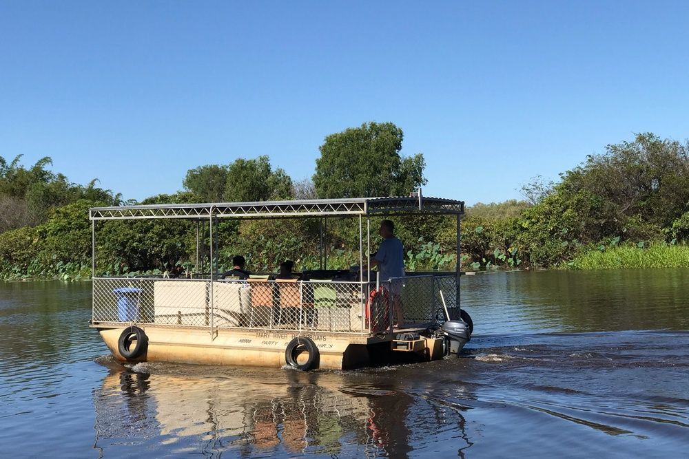 Pontoon boat on water with people, surrounded by lush green trees under a blue sky.