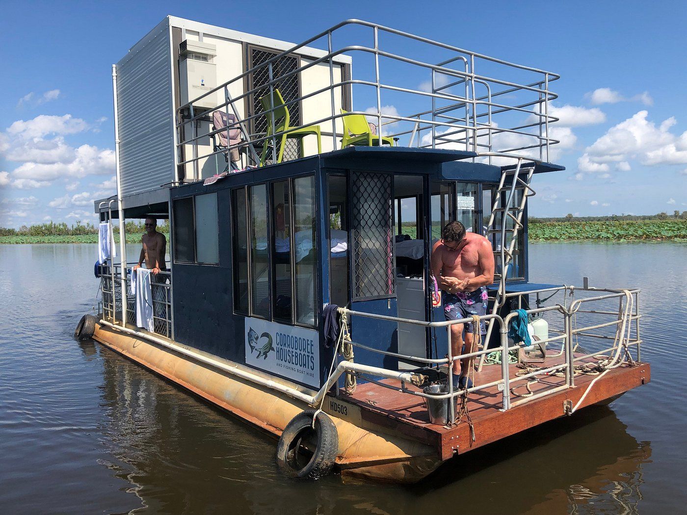 Pink houseboat on water, with white upper structure. A person stands on the side, trees in the background.