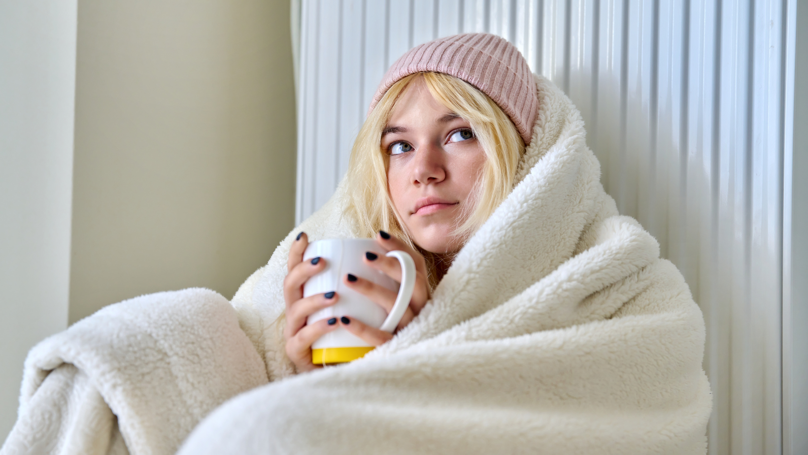 A woman warming herself with a blanket, a beanie, and a cup of coffee.