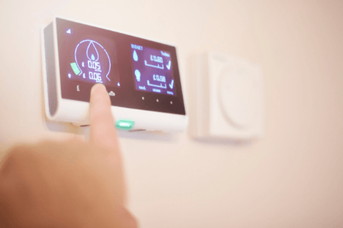 A hand is pointing at a smart thermostat mounted on a wall showing a tracked energy usage on its screen