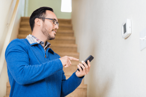 A man wearing a pair of eyeglasses and a blue long-sleeved shirt is standing infront of his smart thermostat while checking its alerts on the smartphone 