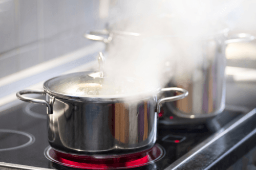 Two stainless pots are on a black electric stove while emitting hot, heavy steam