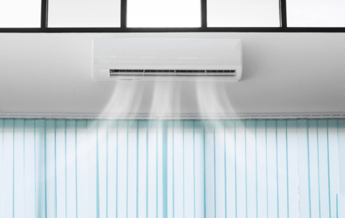 A white mini split ductless air conditioner installed on a ceiling above blue green curtains, showing visible cold air coming out of the unit