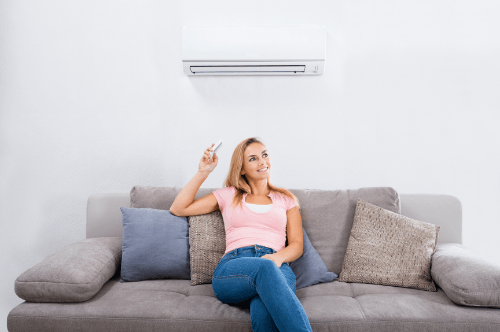 A girl is sitting on the couch while pointing the remote on the white HVAC unit installed up on the wall behind her.