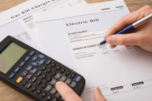 One person's hand is holding a pen over the monthly electric bill while the other hand is typing on a black scientific calculator