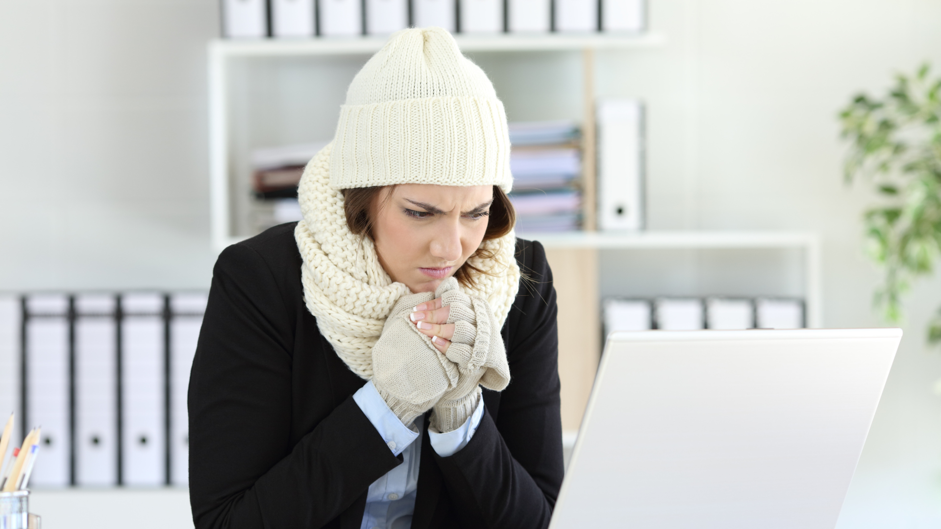 Picture of a woman sitting in front of her laptop inside wearing a scarf, gloves and a hat.