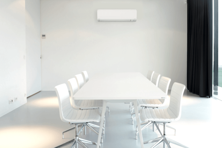 Commercial HVAC is installed on the wall near the end of a long-table. The whole white room is emplty.