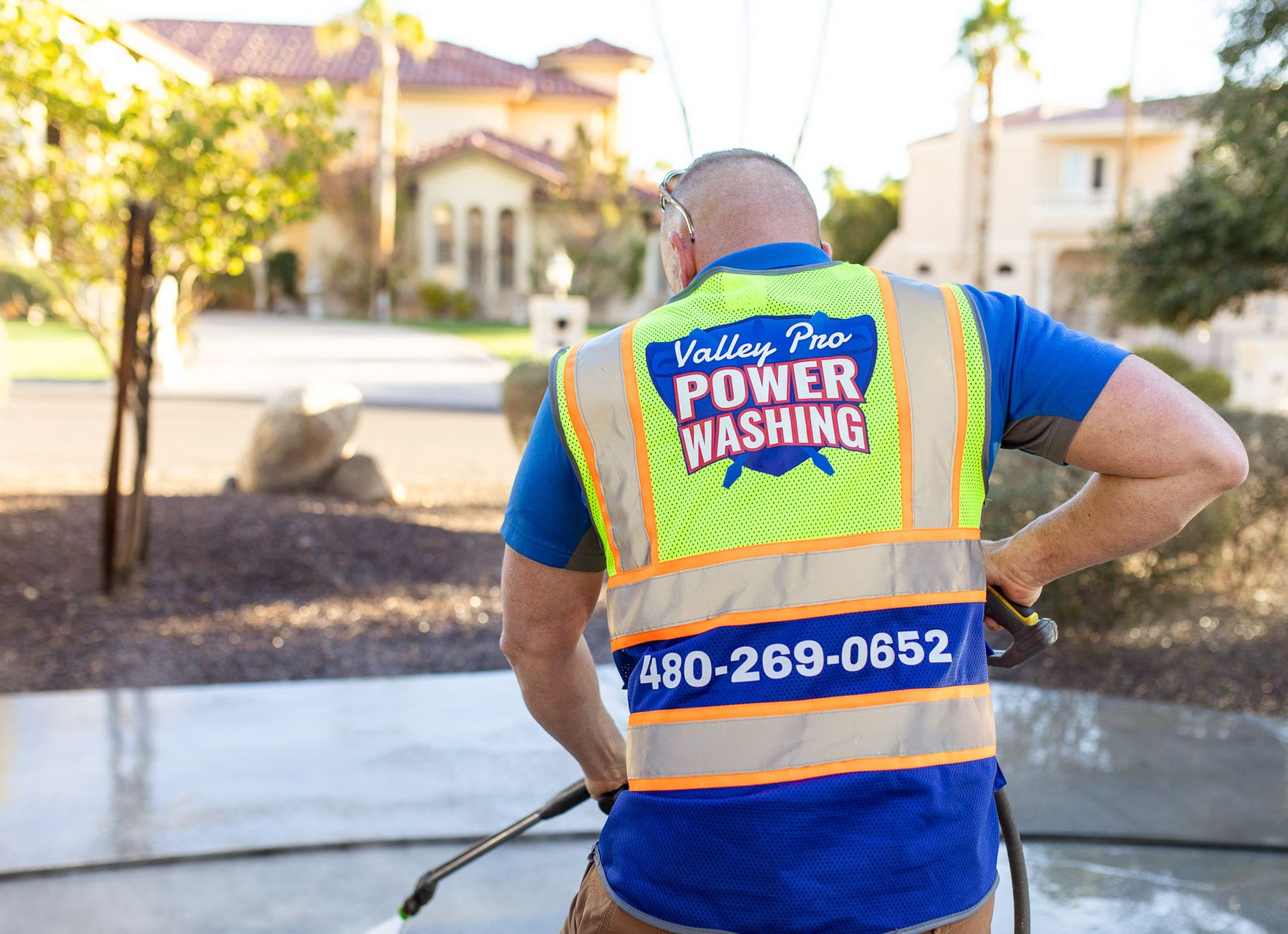Power washing worker in blue shirt and reflective vest holding hose in a sunny neighborhood driveway