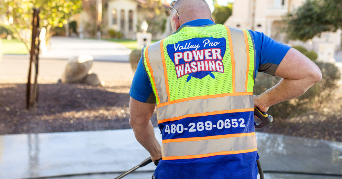 A professional power washer in a high-visibility vest works on a driveway, with company name and phone number visible.