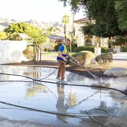A worker in a safety vest uses a circular power washer to clean a wet, reflective concrete driveway outdoors.