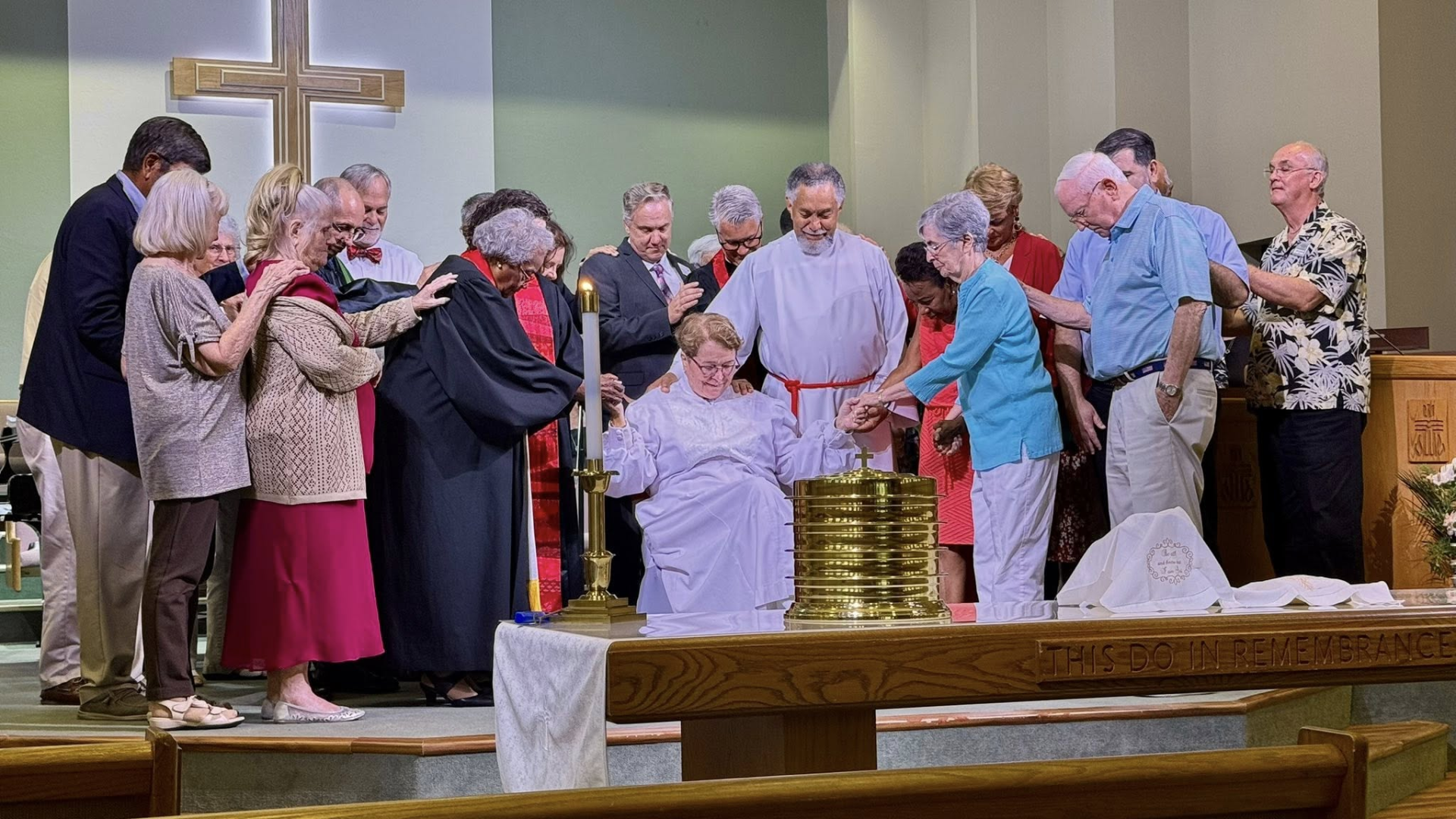 People at a church service, hands on someone sitting near a communion table, cross in background.