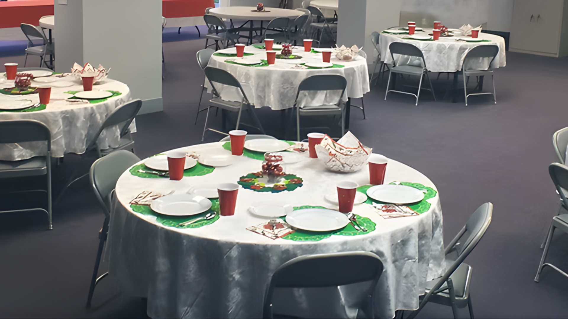 Tables set for a party, covered with white cloths, plates, cups, and decorations. Foldable chairs surround each table.