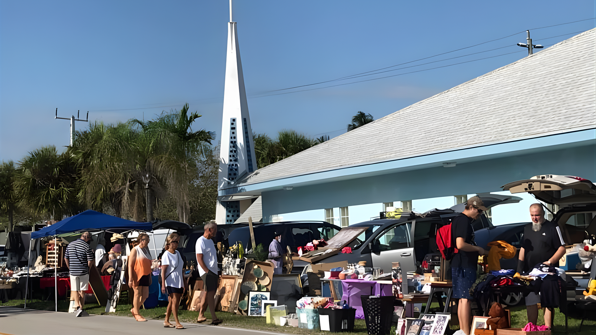 Outdoor church sale with vendors and shoppers on a sunny day. White steeple and light blue building.
