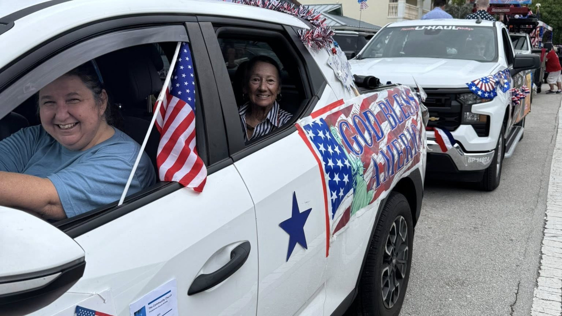 Two women in a decorated white truck, smiling, with American flags, participating in a parade.