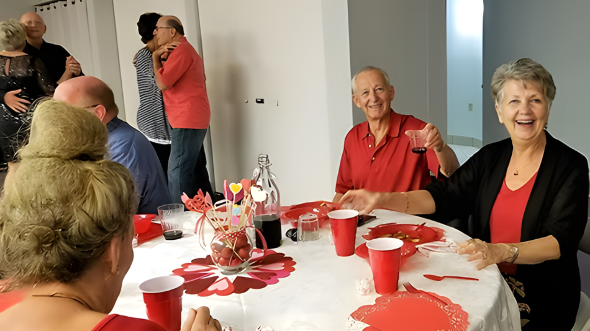 People at a party, some dancing, others at a table. Red, white decor, happy expressions.