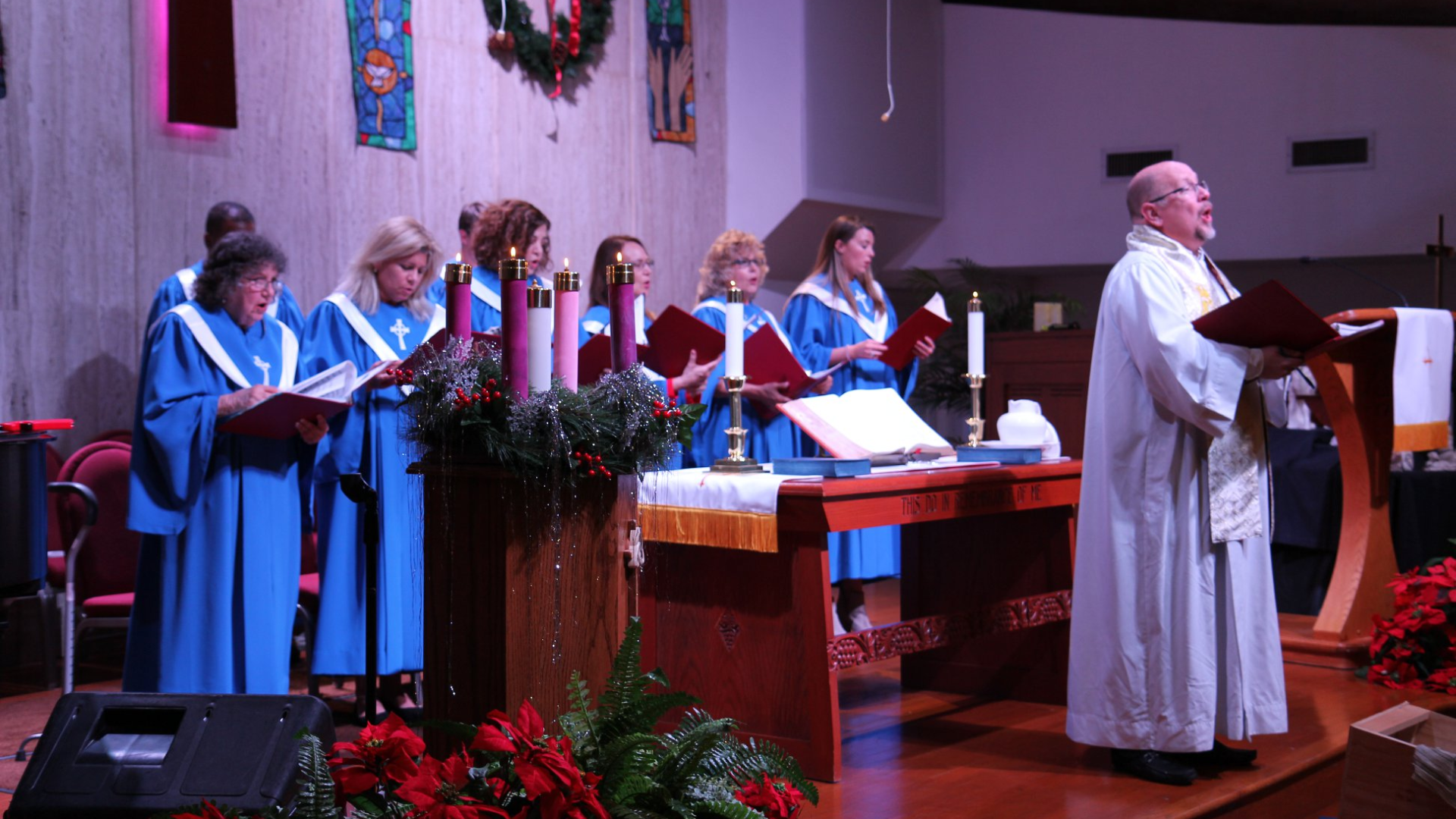 Choir in blue robes sings during a church service; pastor reads from a lectern decorated with Christmas decor.