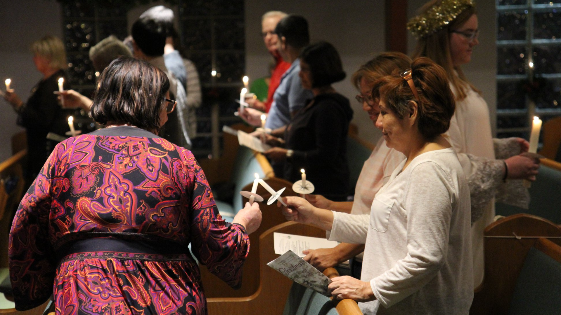 People in a church, holding candles. A candle is lit from another. Dimly lit interior.