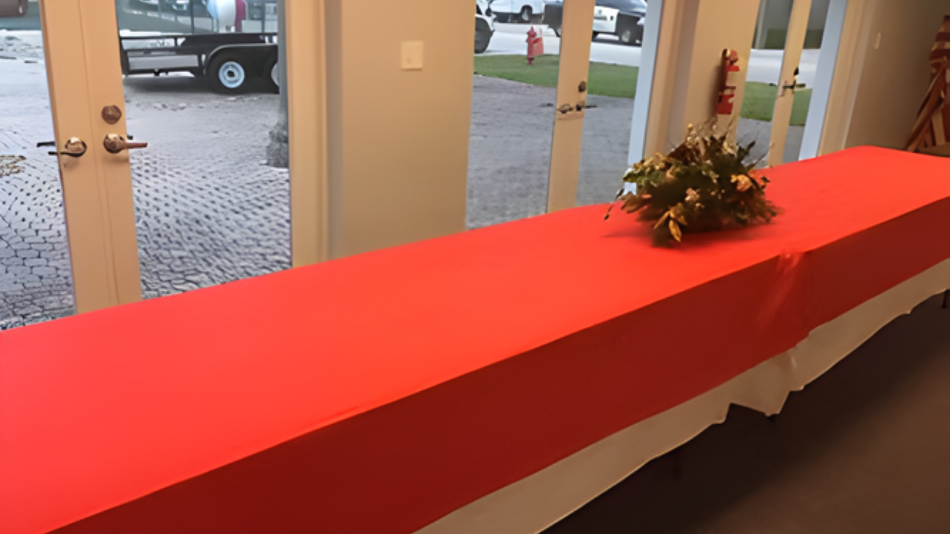 Long red table with floral arrangement, in a room with glass doors, exterior view.